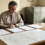 Un conseiller municipal qui déménage doit-il démissionner ? Un conseiller municipal consulte des dossiers vierges et une carte communale sans indications lisibles sur un bureau de mairie, dans une ambiance documentaire réaliste.
