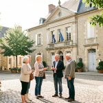 Vue lumineuse d’une mairie française sur une place de village, avec une femme maire et quatre habitants échangeant dans une ambiance conviviale.